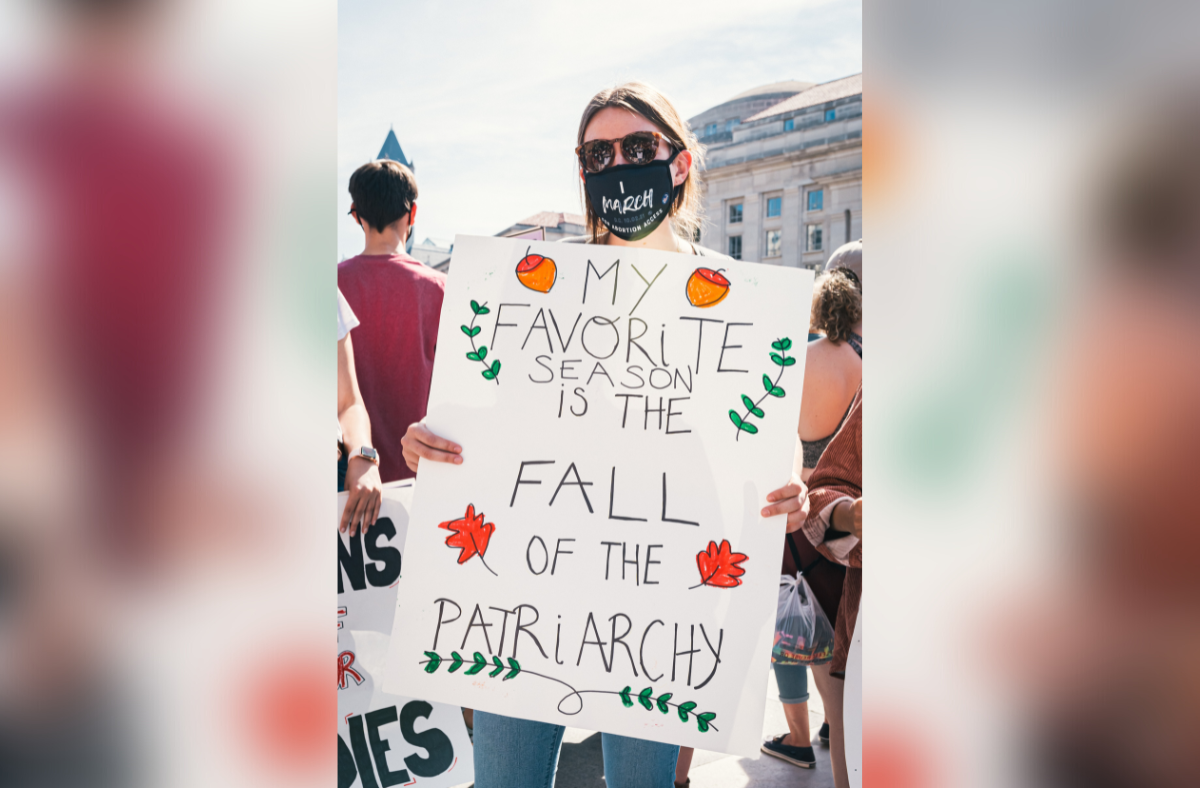 A picture of a person wearing a face mask and sunglasses, participating in a protest, and holding a sign that says: My favourite season is the FALL of the patriarchy